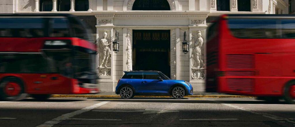 MINI Cooper 3-Türer blau Seitenansicht vor einem historischen Gebäude. Davor und dahinter fahren rote Busse.
