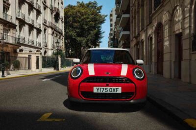 Roter MINI Cooper 3-Türer mit weißen Motorhaubenstreifen, Dach und Spiegelkappen steht in einer Straße mit historischen Gebäuden.