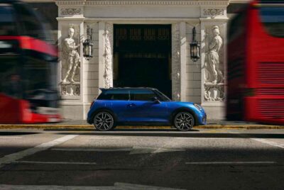 MINI 3-Türer blau Seitenansicht vor einem historischen Gebäude. Davor und dahinter fahren rote Busse.