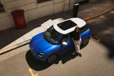 MINI Cooper Electric in blau mit weißem Dach eine Frau lehnt am Fahrzeug, der Blick ist von oben