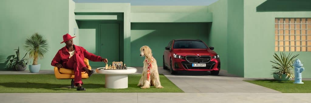 BMW Service mit Proactive Care Mann in roter Kleidung sitzt mit Hund vor einem Haus, ein roter BMW steht vor der Garage