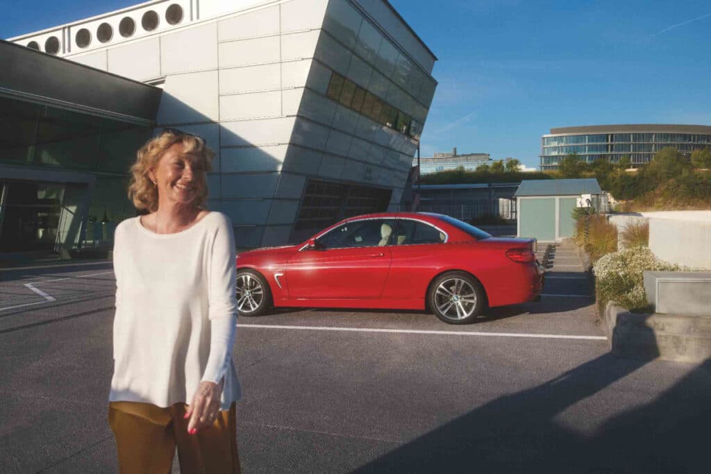 Blonde Frau steht schräg vor einem roten BMW. Im Hintergrund ist eine BMW Werkstatt zu sehen.