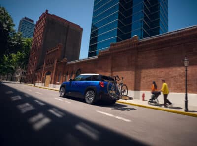 Autohaus Unterberger MINI Aceman in blau mit weißem Dach und Fahrrad auf Fahrradheckträger fährt auf einer Straße - im Hintergrund ist ein rotes Ba
