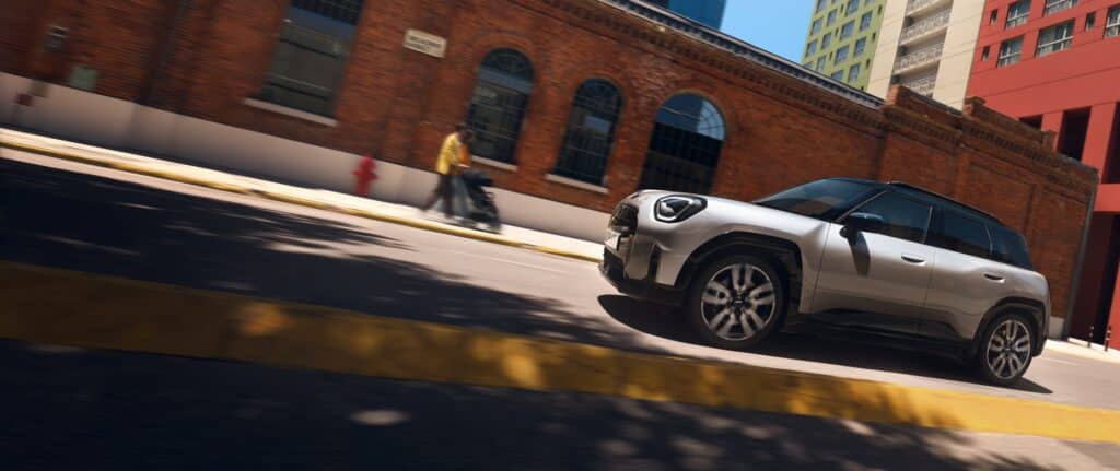 Autohaus Unterberger MINI Aceman in silber fährt auf einer Straße - im Hintergrund ist ein rotes Backsteingebäude