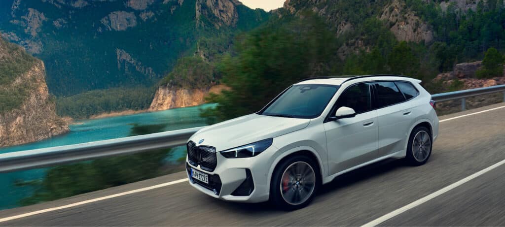 Weißer BMW iX1 fährt auf Straße, im Hintergrund ist ein See zu sehen.