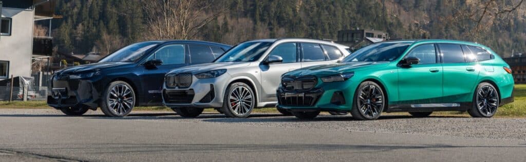 Der BMW iX in schwarz, der BMW iX1 in silber-grau und der BMW i5 Touring in grün in einer Reihe von schräg vorne