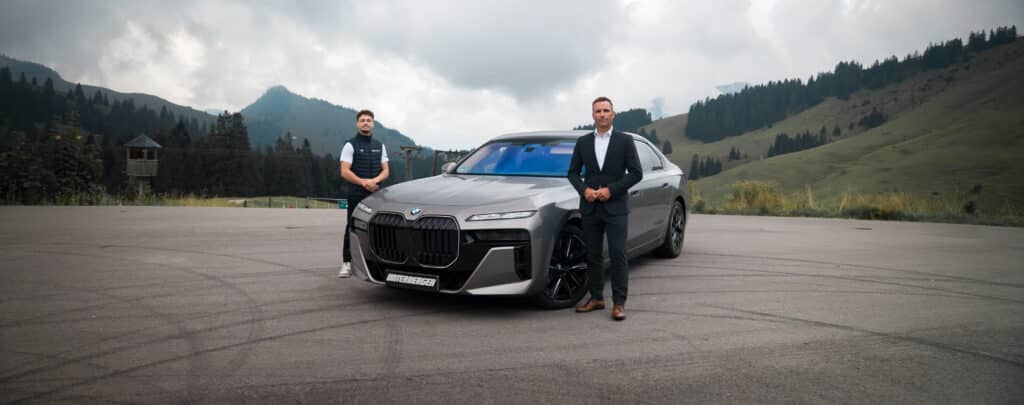 BMW 7er in graumetallic vor bewölktem Himmel und Bergen. Zwei Männer stehen links und rechts neben dem Fahrzeug