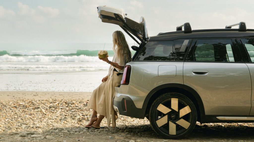 MINI Service Inclusive Frau sitzt auf Ladekante des Kofferraums eines MINI Countryman am Strand. Im Hintergrund ist ein Meer zu sehen.
